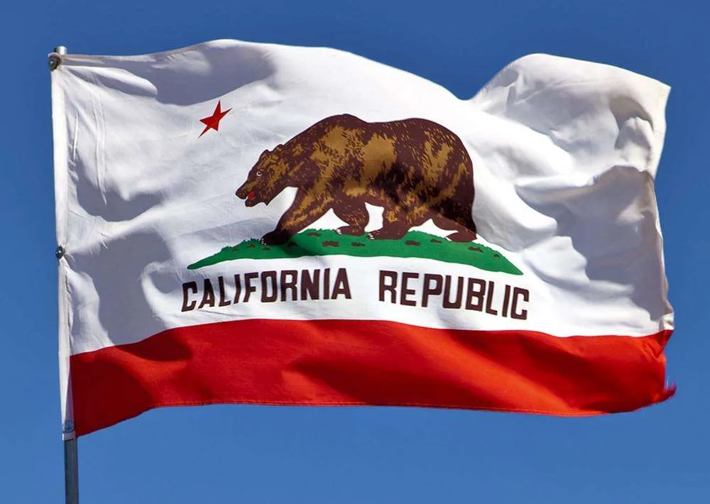 Flag of California (Bear Flag) waving in the breeze, showing the brown bear on a green hill, red star, and white field with 'California Republic' over a red stripe.